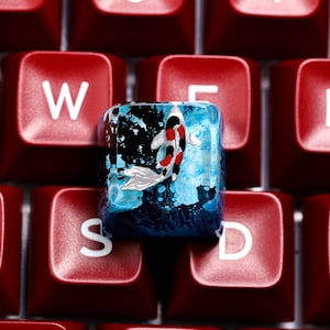 May include: A custom keyboard keycap featuring a koi fish design. The keycap is clear with a blue and black background, and the koi fish has red, black, and white markings. The keycap is set against a background of red keyboard keys.
