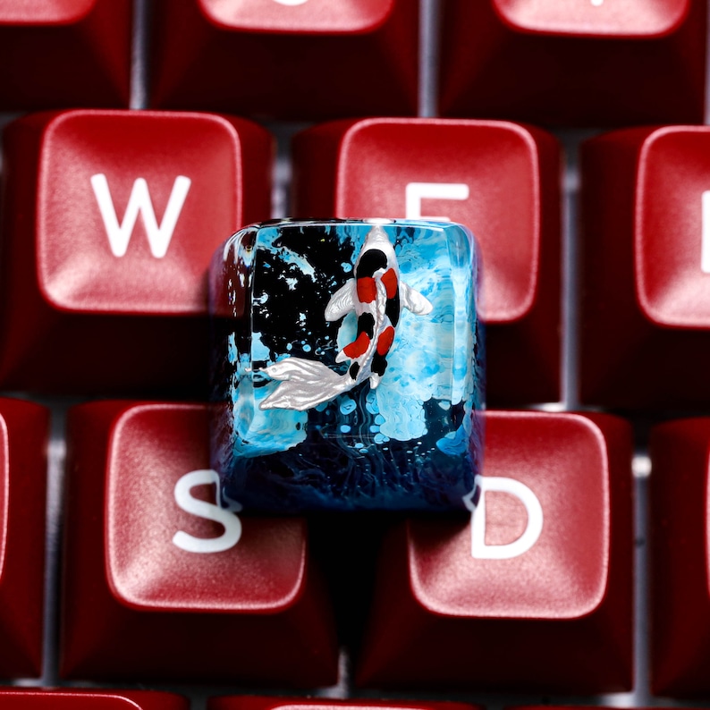 May include: A custom keyboard keycap featuring a koi fish design. The keycap is clear with a blue and black background, and the koi fish has red, black, and white markings. The keycap is set against a background of red keyboard keys.