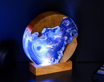 May include: A unique, illuminated resin art piece featuring a swirling blue and white design, reminiscent of a galaxy or deep ocean. The artwork is set within a wooden frame and base, creating a striking contrast. The piece is lit from within, showcasing the intricate details.