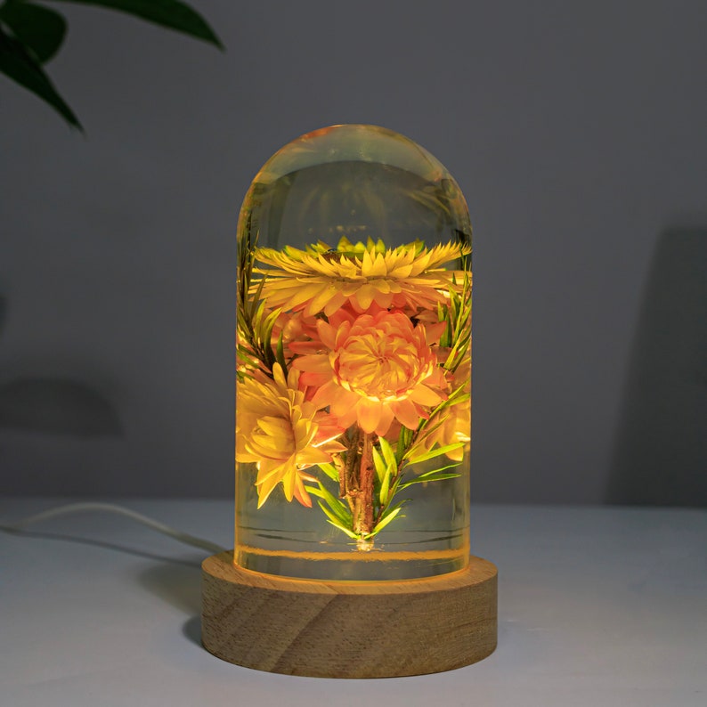 K&ouml;nnte beinhalten: Dekorative Lampe mit getrockneten Blumen in klarem Harz, auf einem Holzsockel. Die Blumen sind gelb und orange. Die Lampe strahlt ein warmes Licht aus, ideal f&uuml;r die Inneneinrichtung. Der Sockel hat ca. 7 cm Durchmesser.