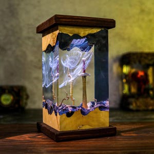 May include: A rectangular wooden lamp with a dark brown top and base. The lamp's core is a clear resin encasing three swords and swirling white and blue smoke-like patterns. The wood has a natural grain and a golden hue.