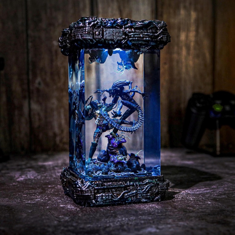 Puede incluir: Una vitrina rectangular e iluminada que presenta una escena de batalla entre un Depredador y un Alien. Las figuras est&aacute;n detalladas en tonos azules y grises, sobre un fondo iluminado en azul. La vitrina tiene una base y una parte superior grises oscuros texturizadas.