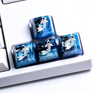 May include: Four custom keyboard keycaps with a koi fish design. The keycaps feature a translucent blue resin base with a white and silver koi fish. The keycaps are square and sit on a white keyboard.