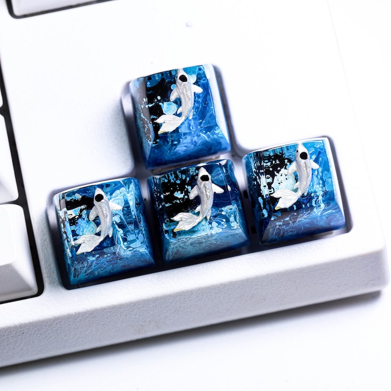 May include: Four custom keyboard keycaps with a koi fish design. The keycaps feature a translucent blue resin base with a white and silver koi fish. The keycaps are square and sit on a white keyboard.