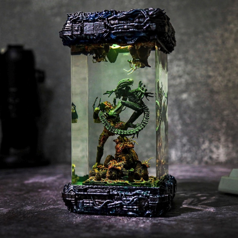 Puede incluir: Escultura rectangular de resina transparente que representa a un Depredador y un Alien en una escena de batalla. La escultura tiene una parte superior e inferior detalladas, de color azul oscuro y negro, con detalles en verde y marr&oacute;n. El interior est&aacute; iluminado en verde.