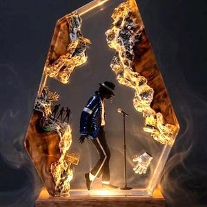 May include: A decorative resin sculpture featuring a figure in a blue sequined jacket, black pants, and a fedora, with a microphone and a single glove. The sculpture is encased in a clear, geometric form with a wooden base and internal rock-like structures.
