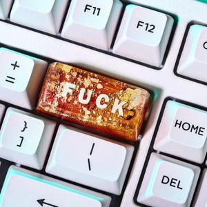 May include: A close-up of a computer keyboard with white keys. The FUCK key is a custom resin keycap with gold flakes and the word "FUCK" in white letters. Other keys include F11, F12, Home, and Del.