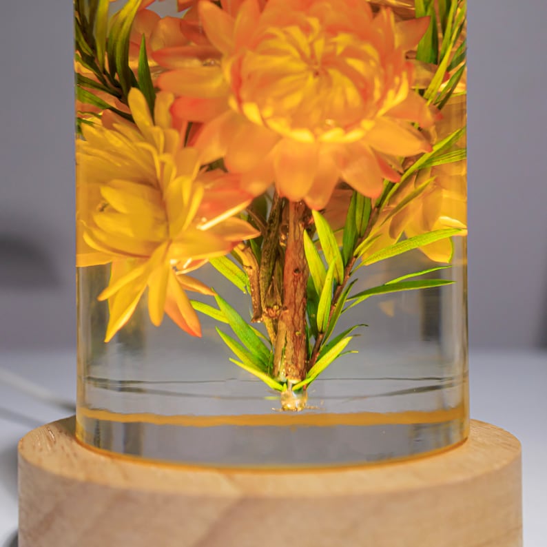 K&ouml;nnte beinhalten: Ein klares, zylindrisches Kunstwerk aus Harz mit getrockneten orangefarbenen Blumen und gr&uuml;nem Laub. Das St&uuml;ck steht auf einem runden, hellen Holzsockel. Die Blumen sind beleuchtet und erzeugen ein warmes Leuchten.