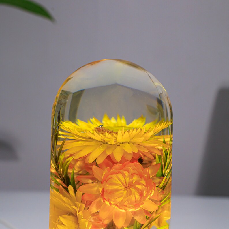K&ouml;nnte beinhalten: Ein klarer, kuppelf&ouml;rmiger Papierbeschwerer aus Harz, der getrocknete gelbe und orangefarbene Blumen umschlie&szlig;t. Die Blumen sind in Schichten angeordnet, mit sichtbaren gr&uuml;nen Stielen und Bl&auml;ttern. Die Oberfl&auml;che ist glatt und poliert.
