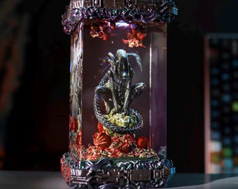 Könnte beinhalten: Rechteckige, beleuchtete Alien-Figur in einer transparenten Box. Der Xenomorph ist dunkelmetallfarben und posiert auf einem grünen Sockel mit roten und orangen Akzenten. Die Box hat einen detaillierten, dunkelgrauen Rahmen.