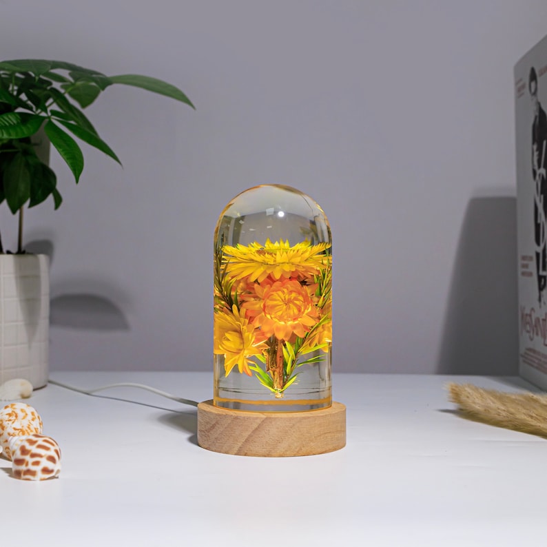 K&ouml;nnte beinhalten: Eine dekorative Lampe mit einem transparenten, kuppelf&ouml;rmigen Glas, das leuchtend gelbe und orangefarbene Blumen umschlie&szlig;t. Die Lampe steht auf einem runden, hellen Holzsockel. Ein wei&szlig;es Kabel erstreckt sich von der Basis.