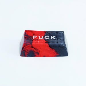 May include: A custom keycap with a red, black, and white marbled design. The word "FUCK" is printed in white across the top of the keycap. The keycap is likely for a mechanical keyboard.