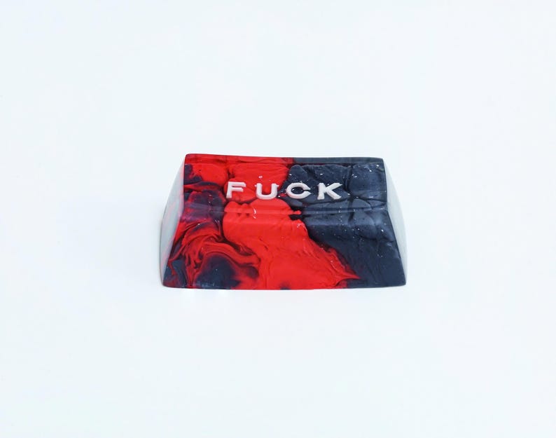 May include: A custom keycap with a red, black, and white marbled design. The word "FUCK" is printed in white across the top of the keycap. The keycap is likely for a mechanical keyboard.