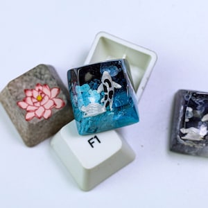 May include: Custom artisan keycaps with unique designs. One keycap features a koi fish swimming in blue resin, another has a pink lotus flower, and others have abstract designs. The keycaps are for a keyboard.
