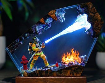 Op de afbeelding: Een decoratieve lamp met een brandweerfiguur in gele en oranje uitrusting, staand naast een rode brandkraan en een klein vuur. De scène is ingesloten in een blok heldere hars met donkere houten randen en straalt een helderblauwe lichtstraal uit.