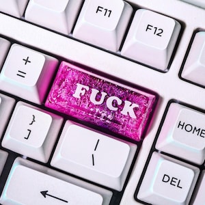 May include: A close-up shot of a computer keyboard with white keys. A custom key in bright pink with the word "FUCK" in white block letters is visible. Other keys include "F11", "F12", "HOME", and "DEL".