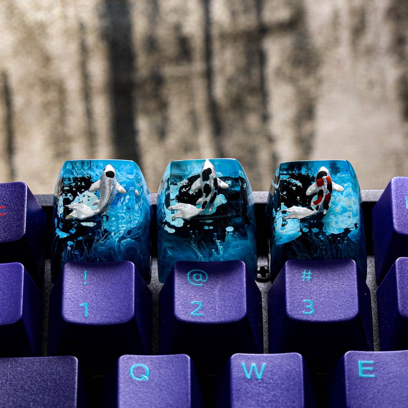 May include: Three custom keyboard keycaps with koi fish designs. Each keycap features a different koi fish, set against a blue and black water-like background. The keycaps are mounted on a purple keyboard.
