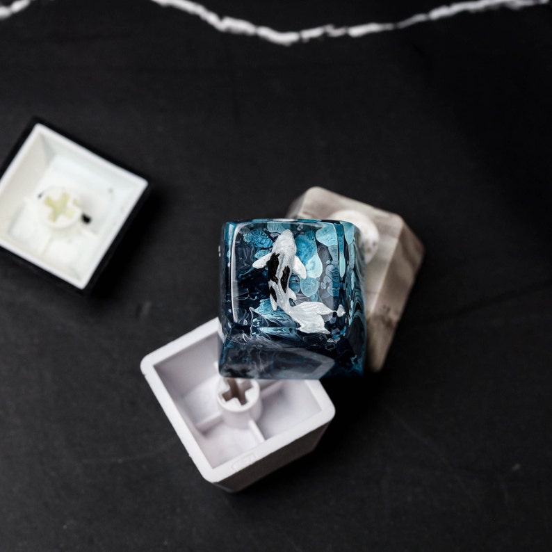 May include: A custom keycap with a koi fish design. The keycap is a translucent blue with white and black koi fish. Two other keycaps are also visible, one white and one with a marble pattern.