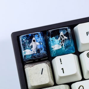 May include: Two custom keycaps with a koi fish design. The keycaps are clear with a blue and white ocean-themed background. One keycap features a white koi fish, and the other has a black and white koi fish. The keycaps are on a black keyboard.