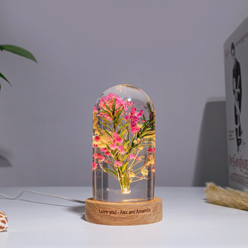 K&ouml;nnte beinhalten: Eine dekorative Glaskuppel mit rosa und gelben Blumen und gr&uuml;nen Bl&auml;ttern, beleuchtet von einem warmen Licht. Die Kuppel steht auf einem Holzsockel mit der Aufschrift "Love you! - Alex and Amanda". Ein wei&szlig;es Kabel geht von der Basis aus.