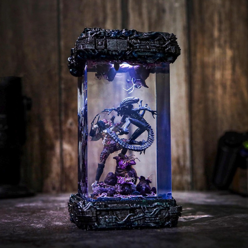 Puede incluir: Diorama detallado de Alien vs. Predator encerrado en una vitrina rectangular transparente. Las figuras se colocan sobre un fondo iluminado en p&uacute;rpura y azul, con una base y parte superior de color gris oscuro detalladas.