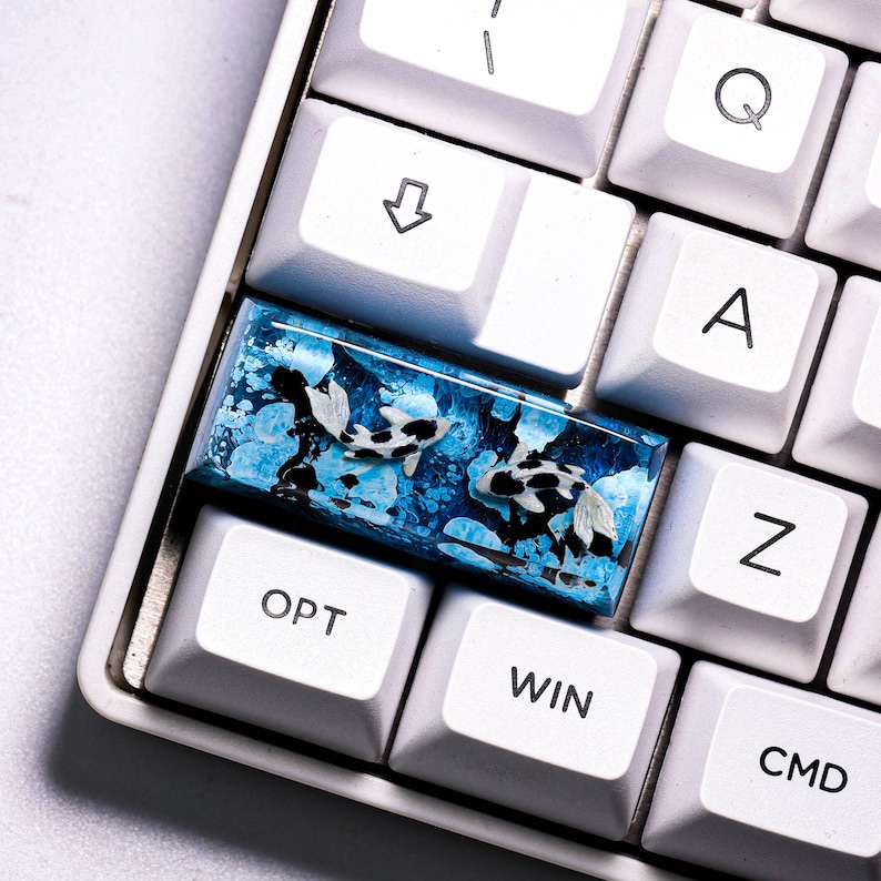 May include: A custom keyboard keycap featuring a koi fish design. The keycap has a rectangular shape with a blue and white background, and two koi fish with black and white markings. The keycap is surrounded by white keyboard keys.