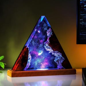 Könnte beinhalten: Eine dreieckige Harzlampe mit Galaxiedesign. Die Lampe hat einen Holzsockel und eine lebendige Darstellung von Sternen, Planeten und einem schwebenden Astronauten. Die Farben sind Blau, Lila und Rot.