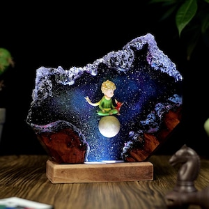 Puede incluir: Luz de noche decorativa con una escultura de resina de una figura sentada con un zorro rojo sobre una luna, sobre un fondo de galaxia estrellada. La escultura está montada sobre una base de madera, creando un brillo ambiental cálido.