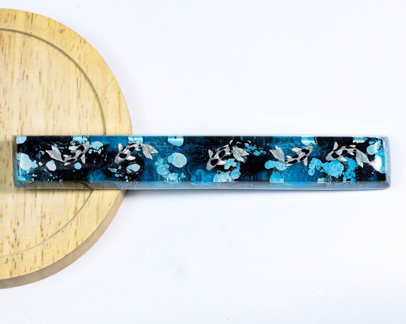 May include: A rectangular resin object with a koi fish design. The design features black and white koi fish swimming in a blue and black water-like background. The object is set on a wooden surface.
