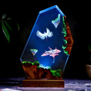 May include: A unique resin art piece featuring a dark blue background with floating islands and two stylized creatures. The artwork is encased in a clear, geometric shape, with a wooden base and accents of green and brown.