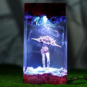 以下が含まれることがあります： 未来的なデザインのミニチュアアニメスタイルのフィギュアを特徴とする長方形の樹脂アート作品。フィギュアは、渦巻く青と白の光の背景に配置され、暗くテクスチャ加工された上部と下部を備えています。