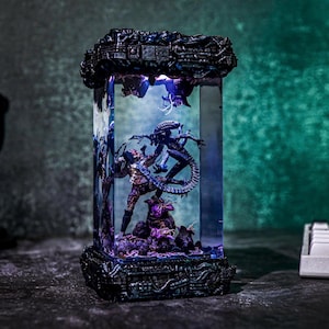Puede incluir: Una vitrina rectangular e iluminada que presenta una escena de batalla entre un Depredador y un Alien. La vitrina tiene una base y una parte superior de color gris oscuro detalladas, con las figuras en su interior. La escena est&aacute; iluminada con tonos azules y morados.