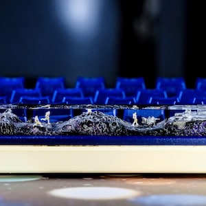 May include: A custom keyboard keycap with a miniature diorama scene. The keycap is clear with a landscape featuring tiny figures, mountains, and a vehicle. The keycap is set on a blue keyboard with a white base.