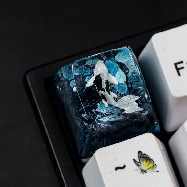 May include: A custom keyboard keycap featuring a koi fish design. The keycap is a translucent blue with a detailed koi fish in black and white. The keycap is surrounded by a dark blue and white swirling pattern.