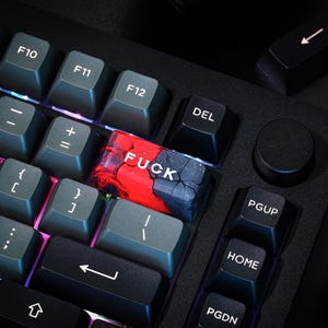 May include: A close-up shot of a black mechanical keyboard with grey keycaps. A custom keycap with the word "FUCK" in white letters is visible. The keycap has a red and blue marbled design. Other keys include function keys and arrow keys.