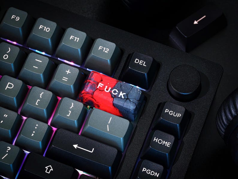 May include: A close-up shot of a black mechanical keyboard with grey keycaps. A custom keycap with the word "FUCK" in white letters is visible. The keycap has a red and blue marbled design. Other keys include function keys and arrow keys.