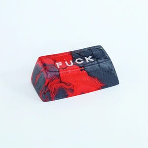 May include: A custom keycap with a red, black, and white marbled design. The word "FUCK" is printed in white, capital letters across the top of the keycap. The keycap is angled and appears to be made of resin.