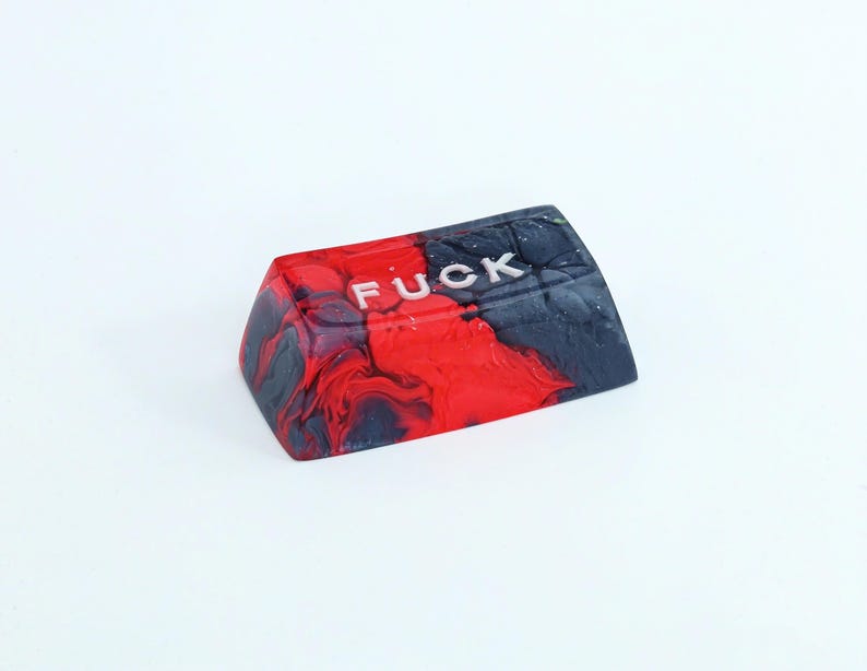 May include: A custom keycap with a red, black, and white marbled design. The word "FUCK" is printed in white, capital letters across the top of the keycap. The keycap is angled and appears to be made of resin.