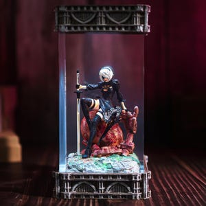 Könnte beinhalten: Detaillierte Figur einer Figur aus dem Spiel Nier: Automata, in einem transparenten Display. Die Figur trägt ein schwarzes Outfit und hält ein Schwert. Der Sockel zeigt eine rote und grüne Landschaft.