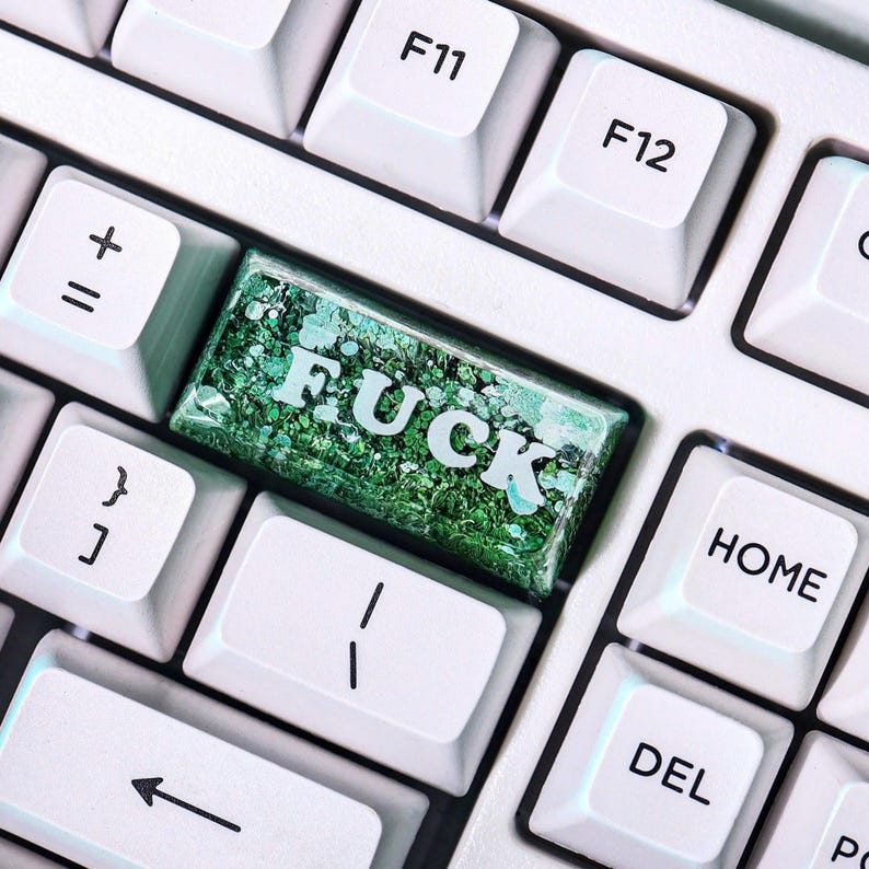 May include: A close-up of a computer keyboard with white keys. The keycap with the word "FUCK" is a custom design with green glitter and white text. Other keys visible include F11, F12, Home, and Del.