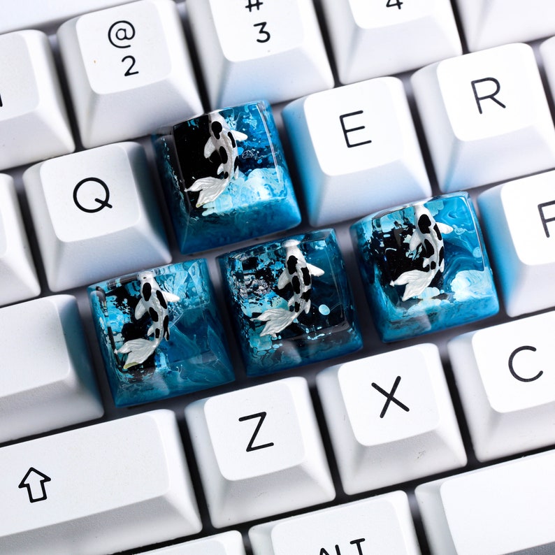 May include: Close-up of a white keyboard with custom keycaps. The keycaps feature a koi fish design in shades of blue, black, and white. The koi fish appear to be swimming in a water-like resin. The keyboard keys are white.