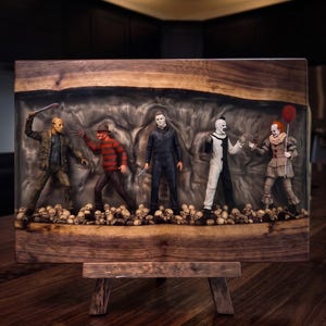 Peut inclure: Un panneau d'exposition rectangulaire en bois à bord brut présentant cinq figurines de personnages d'horreur debout sur un lit de crânes miniatures incrustés dans un centre en résine grise translucide et ondulée. Les figurines incluent un tueur masqué, un personnage en pull rayé aux mains griffues, un homme en combinaison sombre, un clown noir et blanc et un clown tenant un ballon rouge.