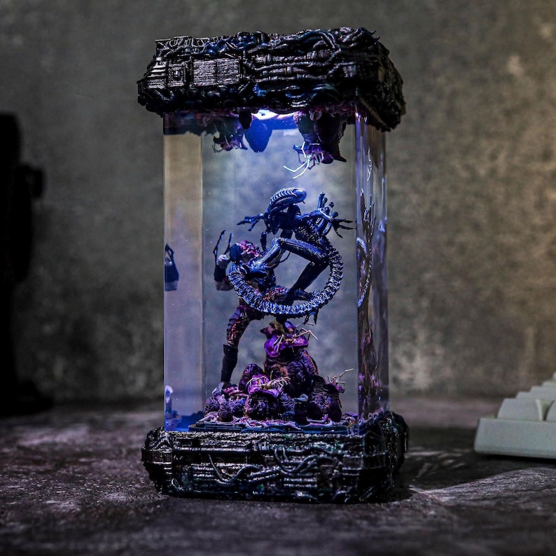 Puede incluir: Una vitrina rectangular iluminada con figuras de Alien y Predator. Las figuras est&aacute;n en una escena de batalla, con el Alien en color azul oscuro y el Predator en color marr&oacute;n rojizo. La vitrina tiene una base y una parte superior negras texturizadas y detalladas.