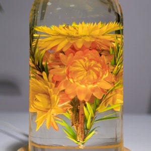 K&ouml;nnte beinhalten: Eine transparente Glasflasche gef&uuml;llt mit leuchtend gelben und orangenen Blumen und gr&uuml;nen Bl&auml;ttern. Die Blumen sind in der Flasche konserviert und bilden eine dekorative Pr&auml;sentation. Die Flasche steht auf einem kleinen, runden Holzsockel.