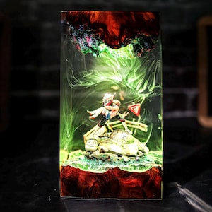 May include: A rectangular resin art piece featuring a miniature figure in action. The piece has a red and brown base, with a green and white swirling design. The top has a dark red and green design. The figure is in the center of the piece.