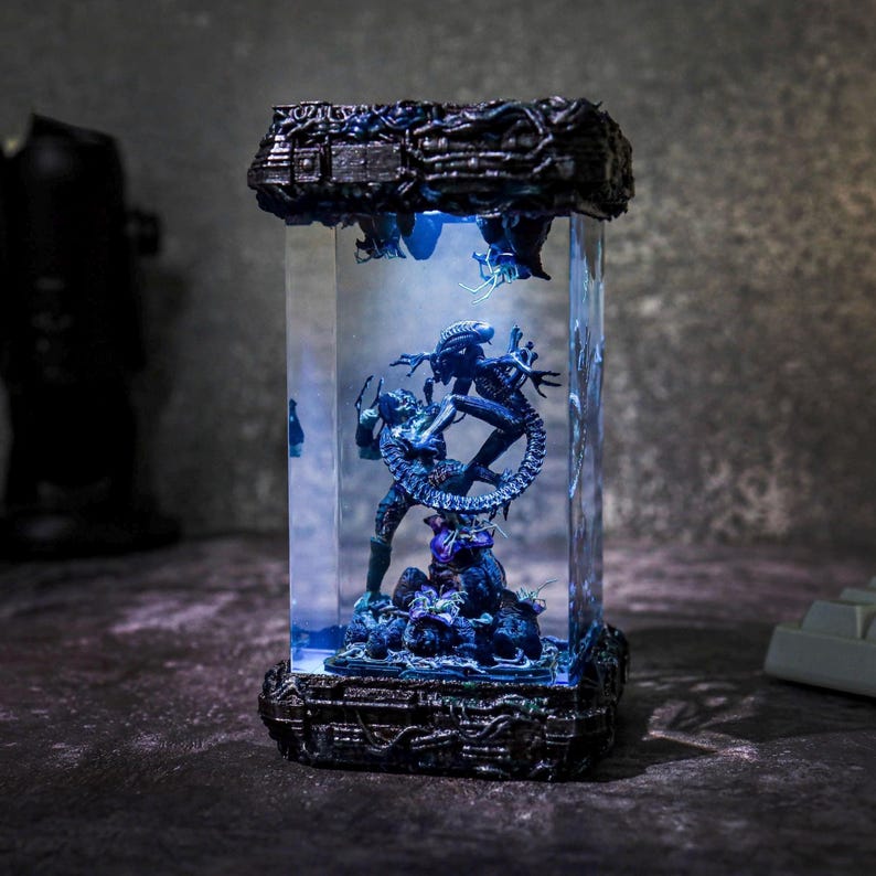 Puede incluir: Una pantalla rectangular iluminada que presenta a un Depredador y un Alien en una escena de batalla. Las figuras est&aacute;n detalladas en tonos azules y negros, sobre un fondo iluminado en azul. La base y la parte superior tienen dise&ntilde;os texturizados intrincados.