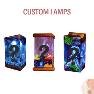 Pode incluir: Três candeeiros personalizados com molduras de madeira e interiores iluminados. Os candeeiros apresentam designs únicos, incluindo uma paisagem urbana, uma cena de fantasia com personagens e uma paisagem iluminada pela lua. O texto "CUSTOM LAMPS" está no topo.