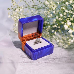 May include: A blue and orange ring box with a diamond ring inside. The box is open, revealing a white interior. The ring has a large, round center stone and smaller stones on the band. White flowers are in the background.