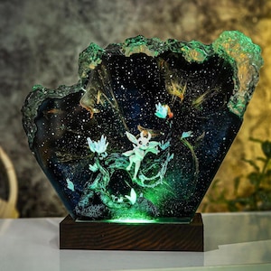 May include: A decorative resin lamp featuring a fantasy scene with a glowing character, flowers, and a starry night sky. The lamp sits on a dark wooden base and has a green, textured border. The interior is illuminated with green light.