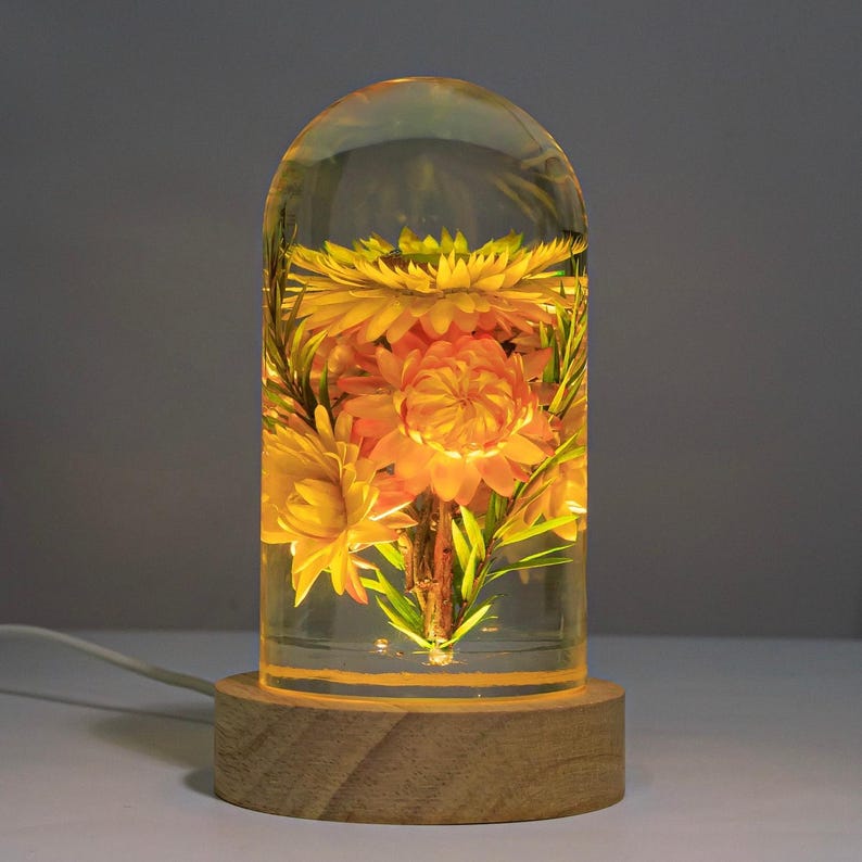 K&ouml;nnte beinhalten: Dekorative Lampe mit einer klaren, gew&ouml;lbten Kunstharzkuppel, die getrocknete gelbe und orangefarbene Blumen umschlie&szlig;t. Die Lampe steht auf einem runden Holzsockel und strahlt ein warmes, gelbes Licht aus. Ein wei&szlig;es Kabel erstreckt sich von der Basis.